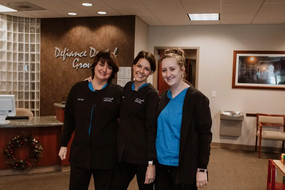 Three Defiance Dental Group staff members at the front desk under the practice's own wall sign